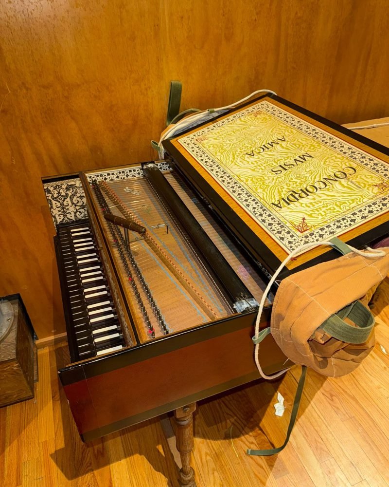 Antique harpsichord with its lid open, showing the keyboard, strings and a decorated soundboard, on a wooden floor