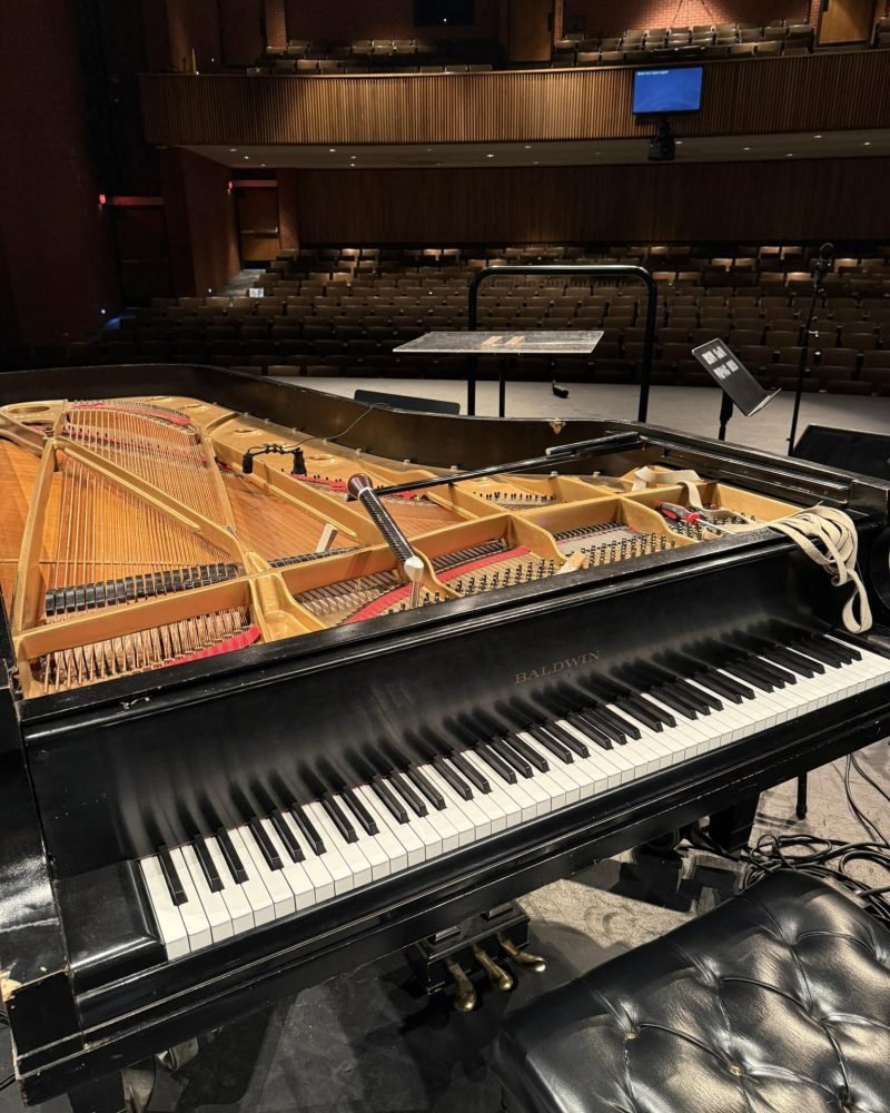 Grand piano with the lid raised and the action exposed on a theater stage, empty seats in the background
