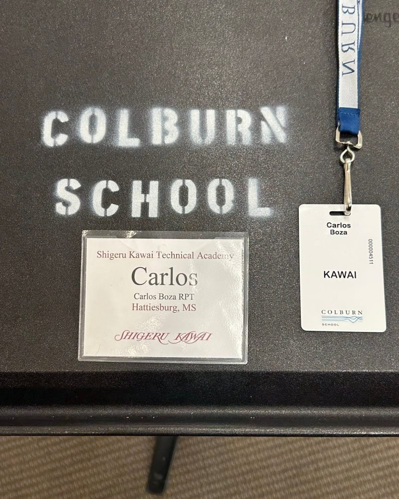 Carlos Boza's name tag from the Shigeru Kawai Technical Academy next to his Kawai lanyard badge, on a Colburn School desk