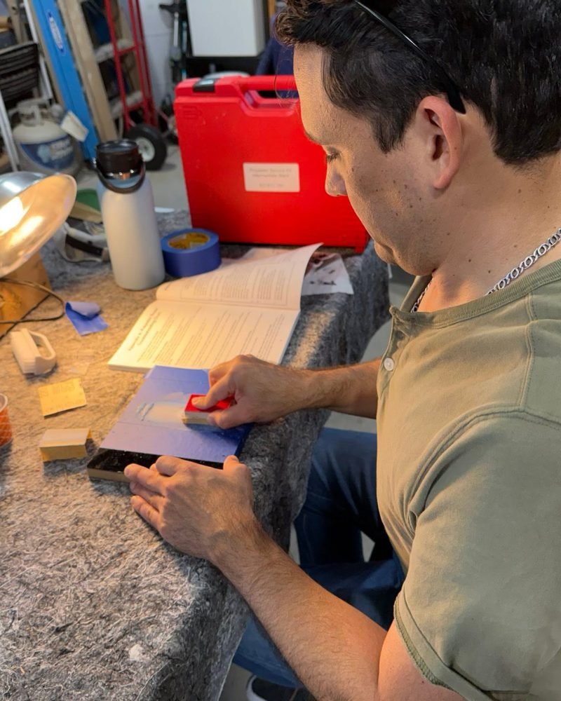 Carlos Boza at a workbench applying a finish touch-up to a black piano part, with a lamp and a reference manual