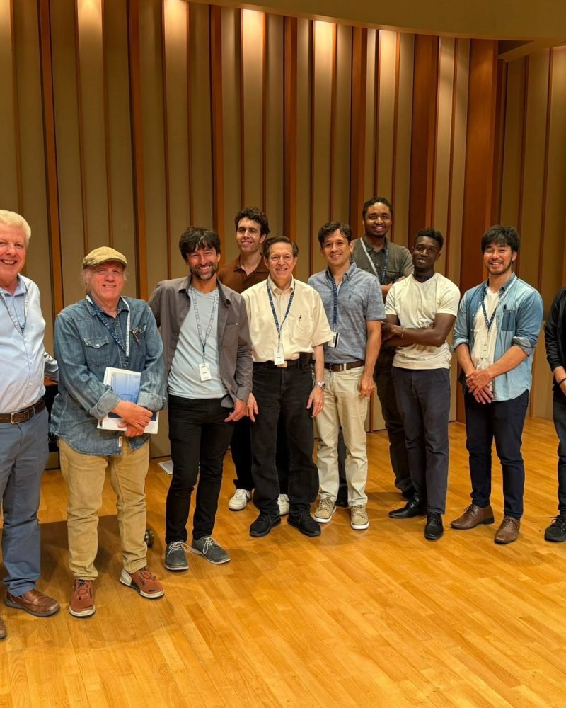 Group of nine piano technicians standing together in a wood-paneled recital hall after a training course