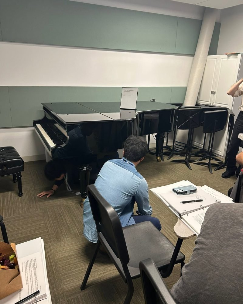 Technician kneeling under a grand piano in a practice room, examining the instrument from below