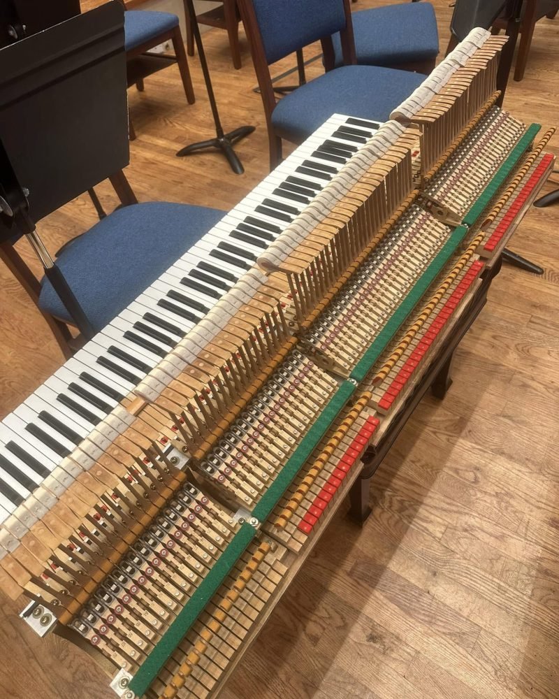Upright piano action removed and laid on its side on the floor, hammers and keys exposed, chairs in the background